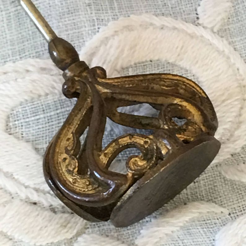 May include: A gold-toned metal pin with a decorative, ornate design. The pin has a long, thin stem and a large, intricate head.