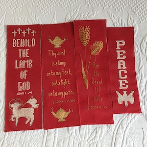 Op de afbeelding: Vier rechthoekige bladwijzers van rode stof met kruissteekborduurwerk. De ontwerpen bevatten een lam met de tekst Behold the Lamb of God John 1-29, een lamp met Thy word is a lamp unto my feet, korenaren en het woord Peace.