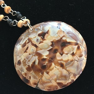 May include: A round pendant necklace with a mosaic-like design in shades of brown and cream. The pendant is attached to a chain with small wooden and black beads.