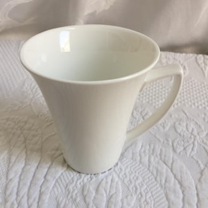 May include: A white ceramic mug with a flared top and a curved handle. The mug is set against a white, textured background. The mug's design is simple and elegant, suitable for coffee or tea.