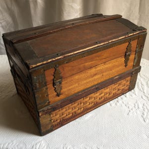 Puede incluir: Un peque&ntilde;o cofre de madera con herrajes de lat&oacute;n y un patr&oacute;n decorativo en la parte inferior. El cofre tiene una tapa con bisagras y est&aacute; hecho de madera con un acabado natural.