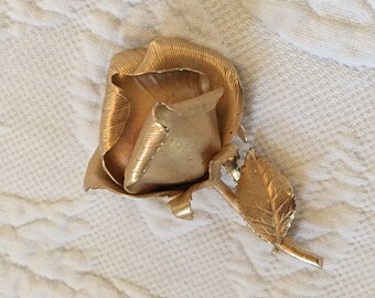 Könnte beinhalten: Eine goldfarbene Rosenbrosche mit einem einzelnen Blatt. Die Rose ist aus Metall und hat eine strukturierte Oberfläche.