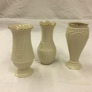 May include: Three cream ceramic vases with gold rims. The vases have varied shapes and textures, including a dotted pattern and vertical lines. The vases are decorative and suitable for displaying flowers.