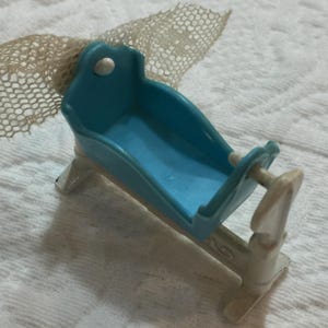Może przedstawiać: Miniaturowa niebieska kołyska z białymi podporami i dekoracyjną siatką. Kołyska jest wykonana z tworzywa sztucznego i ma zakrzywiony kształt. Kołyska to zabawka w stylu vintage.