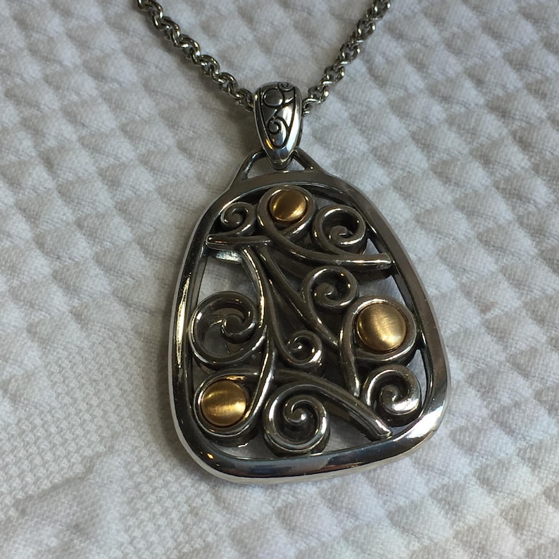 May include: A silver-toned pendant necklace featuring an openwork design with swirling metal scrolls and three circular gold-toned accents. The pendant is suspended from a thick silver-toned chain with a decorative bail at the top of the piece.