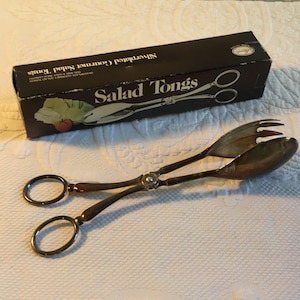 Puede incluir: Pinzas de ensalada gourmet plateadas, de aproximadamente 26,7 cm de longitud, junto a una caja negra con las palabras "Salad Tongs" y "Silverplated Gourmet Salad Tongs". Las pinzas tienen un mango en forma de tijera y un extremo de tenedor y cuchara.