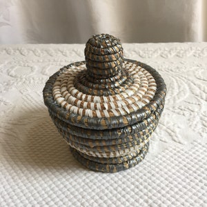 以下が含まれることがあります： 蓋付きの小さな丸い籐のバスケット。バスケットは自然素材で作られており、グレーとホワイトのストライプ模様になっています。蓋は丸みを帯びています。