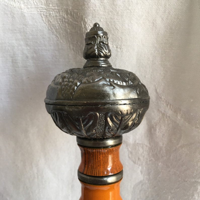 Puede incluir: Primer plano de una empu&ntilde;adura de espada decorativa de color plateado con intrincados dise&ntilde;os florales. La empu&ntilde;adura est&aacute; unida a un eje de madera con un patr&oacute;n de rayas marrones y naranjas.