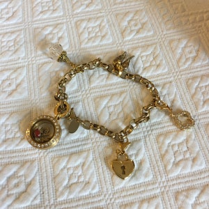 May include: A gold charm bracelet with a variety of charms, including a locket, a heart-shaped padlock, a key, and a winged angel. The charms are attached to a gold chain.