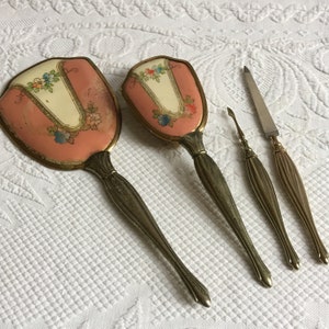 Op de afbeelding: Een vintage set van vier zilverkleurige toiletartikelen. De set bevat twee handspiegels met roze en witte bloemenmotieven, een nagelvijl en een nagelriemduwer.