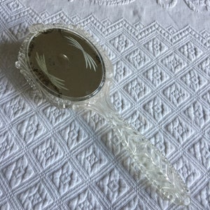May include: A clear plastic hand mirror with an oval-shaped mirror and a decorative handle. The mirror has a etched design of leaves on the surface.