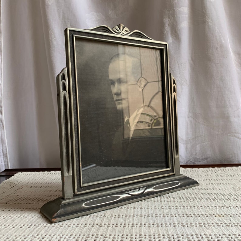 May include: A silver-toned picture frame with a black mat and a black and white photograph of a man. The frame has a decorative design and stands on a base.