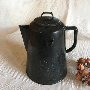 May include: A black enamel coffee pot with a lid and handle. The pot has a spout for pouring and a speckled white pattern. The coffee pot is sitting on a white, textured surface.