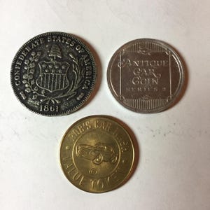 Może przedstawiać: Wyświetlane są trzy monety. Moneta w lewym górnym rogu jest srebrna i zawiera napis "CONFEDERATE STATES OF AMERICA 1861". Moneta w prawym górnym rogu jest srebrna i zawiera napis "ANTIQUE GAR GOIN SERIES 2". Dolna moneta jest złota i zawiera napis "ROB'S CAR WASH VALUE TOKEN".