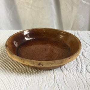 May include: A round, shallow ceramic bowl with a glossy, dark brown glazed interior and a contrasting matte, light tan exterior rim. The bowl features visible concentric circular markings on the base and a smooth, slightly uneven texture throughout.