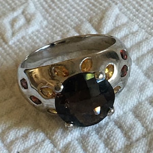 May include: A silver ring with a large, dark brown gemstone surrounded by smaller, yellow and red gemstones.