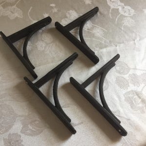 May include: Four black metal shelf brackets. Each bracket has a flat base and a curved support arm. The brackets are designed to be mounted on a wall to support shelves. The brackets are made of a dark, possibly cast iron, material.