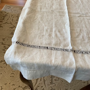 Può includere: Un tovaglia di lino bianco con un delicato bordo all'uncinetto. La tovaglia è drappeggiata su un tavolo e ha un sottile motivo testurizzato.