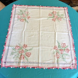 Puede incluir: Mantel blanco con un borde de ganchillo en rosa y verde. El mantel tiene diseños florales bordados con tulipes rosas y verdes.