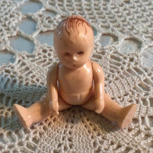 May include: A small, vintage plastic doll in a peach hue. The doll has jointed arms and legs, and brown painted hair. The doll is seated with its legs apart. The doll is a collectible toy, reminiscent of childhood.