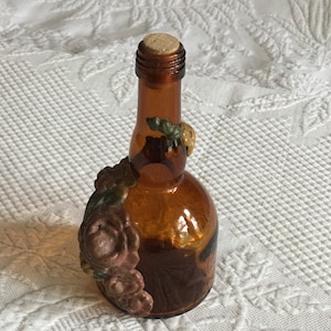 Puede incluir: Una pequeña botella de vidrio ámbar con un tapón de corcho. La botella está decorada con un diseño floral en tonos marrones, verdes y amarillos.
