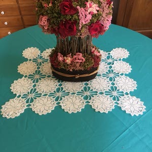 May include: A white, crocheted lace doily with a floral design sits on a teal tablecloth. A decorative floral arrangement with red roses and pink flowers is centered on the doily. The arrangement is in a dark brown container.