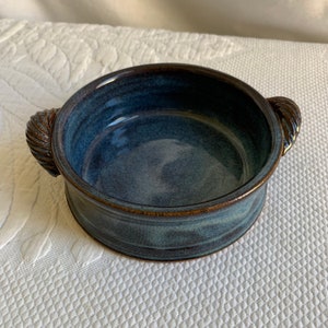 Könnte beinhalten: Eine kleine, blaue Keramikschale mit zwei Schlaufehenkeln. Die Schale hat eine gesprenkelte Glasur und einen braunen Rand.