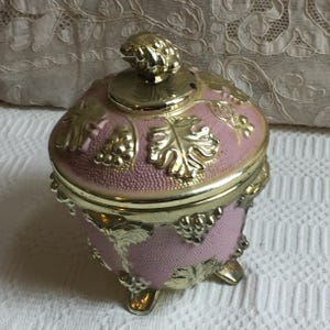 May include: A pink trinket box with gold-coloured accents. The box has a textured surface and is decorated with gold grape and leaf designs. It has a matching lid with a decorative knob and rests on three small feet.