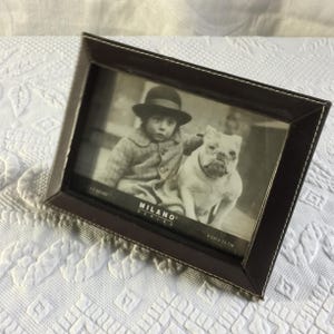 May include: A dark brown picture frame with white stitching around the edges. The frame holds a black and white photograph of a child in a hat and a bulldog. The frame is labeled "MILANO SERIES" and is 9 cm x 13 cm.
