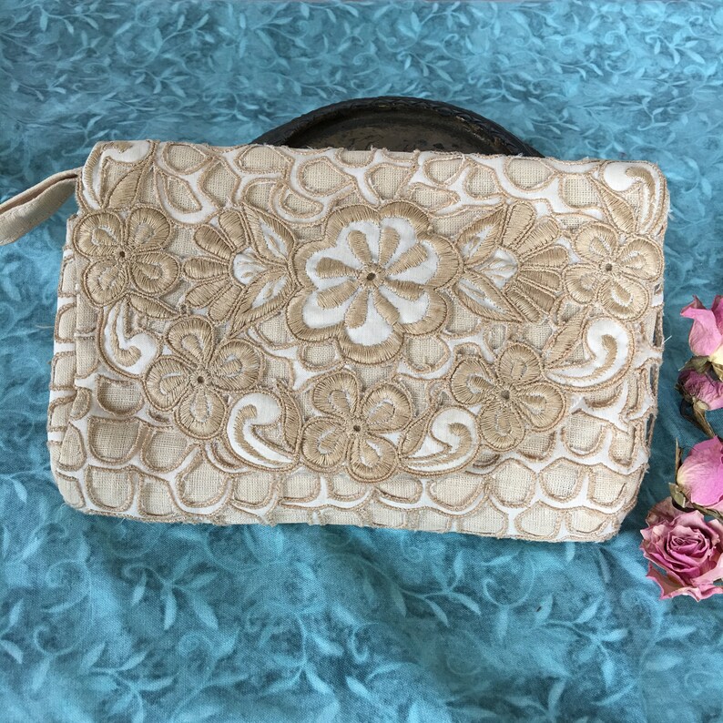 May include: A white clutch purse with a floral design embroidered in beige thread. The purse has a simple, elegant design.