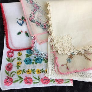 May include: A collection of vintage handkerchiefs. One features a red crocheted edge and a holly and bell design. Another has a pink floral border. A third has a cream-colored lace edge. The bottom handkerchief has pink tulips and blue roses.