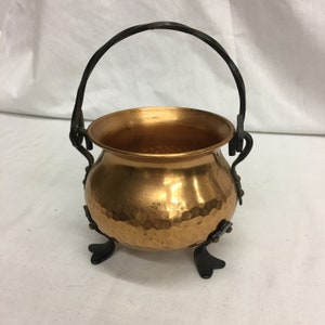 May include: A small copper pot with a hammered finish and a black wrought iron handle and legs.