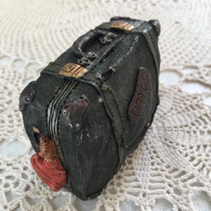 Pode incluir: Uma mala preta em miniatura com detalhes dourados, alças e uma pega. A mala tem uma superfície texturizada e uma etiqueta de couro castanho que diz "BOYDS". Um elemento decorativo vermelho e castanho está preso na lateral.