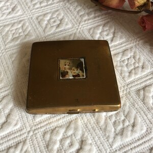 Op de afbeelding: Een vintage, vierkant compact poederdoosje in messingkleur. Het compacte poederdoosje heeft een kleine, rechthoekige, kleurrijke afbeelding van een vrouw en kinderen. Het compacte poederdoosje is gesloten en ligt op een wit, patroon oppervlak.