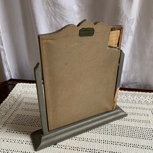 May include: A gray wooden vanity mirror with a decorative top and a stand. The mirror has a label on the back that reads "Larkin Photo Supply Co. - 444 S. Franklin St. - Chicago, Ill. - U.S.A."
