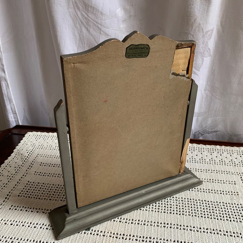 May include: A gray wooden vanity mirror with a decorative top and a stand. The mirror has a label on the back that reads "Larkin Photo Supply Co. - 444 S. Franklin St. - Chicago, Ill. - U.S.A."