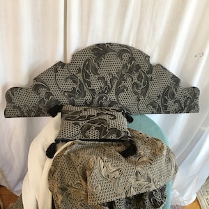 May include: A black and gold patterned headboard with a decorative arched top. The headboard is upholstered in a fabric with a floral design. A black and gold patterned pillow is placed on top of the headboard.