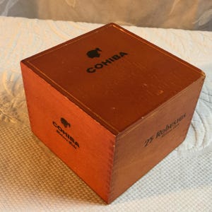 Puede incluir: Una caja de madera de color marrón rojizo. La palabra "COHIBA" está impresa en negro en la parte superior y lateral. La caja también tiene el texto "25 Robustos" impreso en el lateral.