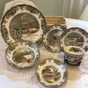 Może przedstawiać: Kolekcja zabytkowej zastawy stołowej ceramicznej przedstawiającej zimowy krajobraz z czerwonym budynkiem. Zestaw zawiera talerze, miski i filiżankę do herbaty, wszystkie z ozdobnym liściastym obramowaniem. Talerze mają średnicę około 25 cm.