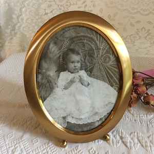May include: An oval-shaped brass picture frame with a black and white photo of a baby girl wearing a white dress. The frame has a decorative design and stands on three feet.