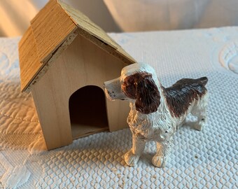 May include: A brown and white toy dog standing next to a small wooden dog house. The dog house has a simple design with a pitched roof.