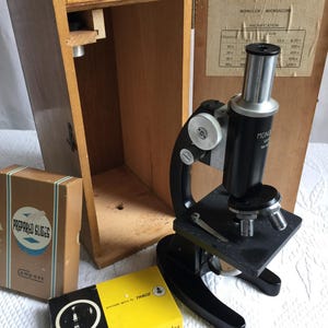 May include: A black Monolux microscope with a silver eyepiece and objective lenses, positioned next to its open wooden storage case. Included are two rectangular boxes labeled Prepared Slides and microscope slides by Tasco.