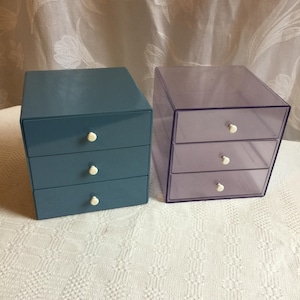 May include: Two small drawer chests. One is teal with three drawers and white knobs. The other is transparent purple with three drawers and white knobs. Both are cube-shaped and sit on a white, textured surface.