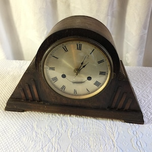 May include: A dark brown wooden mantel clock with a rounded top and a triangular base. The clock face is circular with a metallic finish, black Roman numerals, and two black hands. The wooden frame features decorative vertical grooved accents.