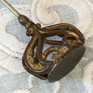 May include: A gold-toned metal pin with a decorative, ornate design. The pin has a long, thin stem and a round base.