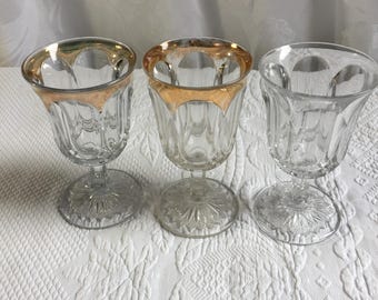 Vintage 3 Heisey Wassergläser auf Fuß, zwei mit goldenem oberen Rand. Verziert mit Zierelementen und einem sonnenförmigen Bodenmuster.