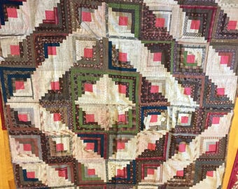 May include: A handmade quilt with a geometric pattern. The quilt features squares with a central pink square surrounded by various fabrics in shades of brown, green, blue, and cream. The quilt is likely made of cotton or a similar fabric.