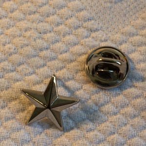 Op de afbeelding: Een zilverkleurige ster-vormige reversspeld en een bijpassende pin-achterkant. De pin heeft een vijf-puntige ster ontwerp en een glanzende, metallic afwerking. De pin-achterkant is een kleine, ronde schijf met een pin om de pin vast te zetten.