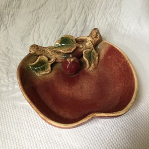 May include: A ceramic apple-shaped dish with a raised, three-dimensional design of an apple branch with leaves and a small apple. The dish is glazed in shades of red, brown, and green.