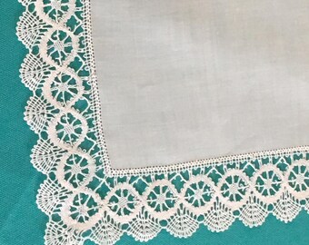 May include: A white handkerchief with a delicate, scalloped lace border. The lace features a repeating floral pattern. The handkerchief is made of a fine, sheer fabric, and is displayed on a teal background.